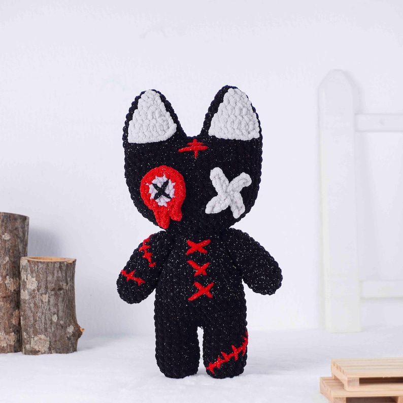 May include: A handmade black crochet doll with white ears and facial details. The doll has red stitched accents and a red and white eye design. The doll is standing upright on a white surface.