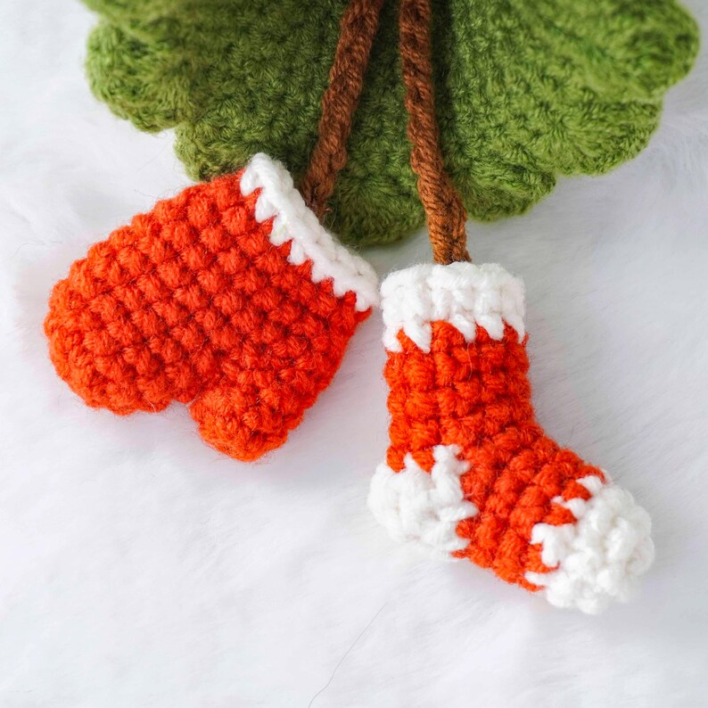 Op de afbeelding: Gehaakte kerstversieringen met een paar oranje wanten en een sok met witte rand. De ornamenten zijn bevestigd aan een groene gehaakte boom met bruine stelen. De wanten en de sok hebben witte accenten.