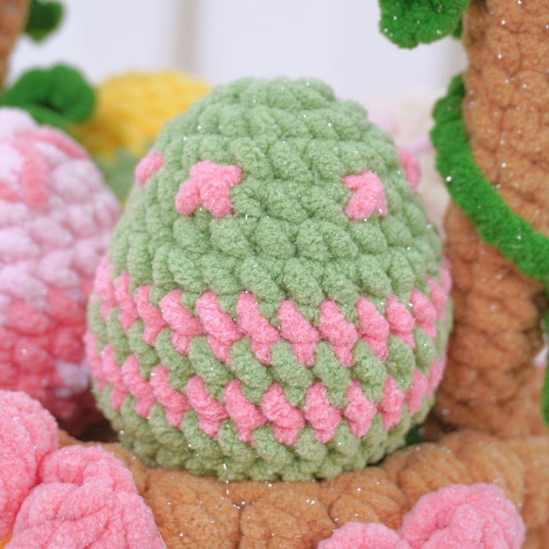 May include: A green crocheted Easter egg with pink accents and glitter. The egg is surrounded by other crocheted Easter eggs in various colors.