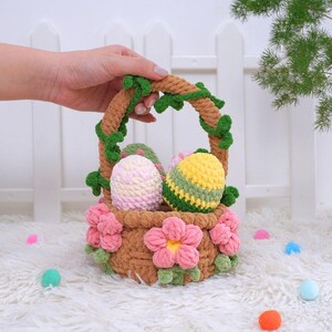 May include: A brown crocheted basket with a green vine handle and pink flowers. The basket is filled with three crocheted Easter eggs, one pink and white, one yellow and green, and one green and yellow.