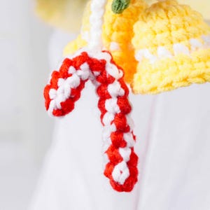 May include: Close-up of a handmade crochet ornament featuring a red and white candy cane shape. The ornament is attached to a yellow bell-shaped crochet piece with white accents. A small green crochet piece is also visible.