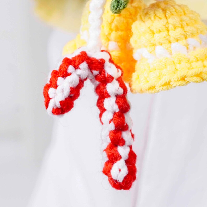 May include: Close-up of a handmade crochet ornament featuring a red and white candy cane shape. The ornament is attached to a yellow bell-shaped crochet piece with white accents. A small green crochet piece is also visible.