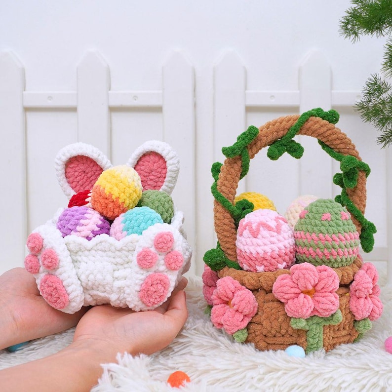 May include: Two crocheted Easter baskets filled with colorful yarn eggs. One basket is shaped like a bunny with pink ears and paws. The other basket is a brown woven basket with pink flowers and green vines.