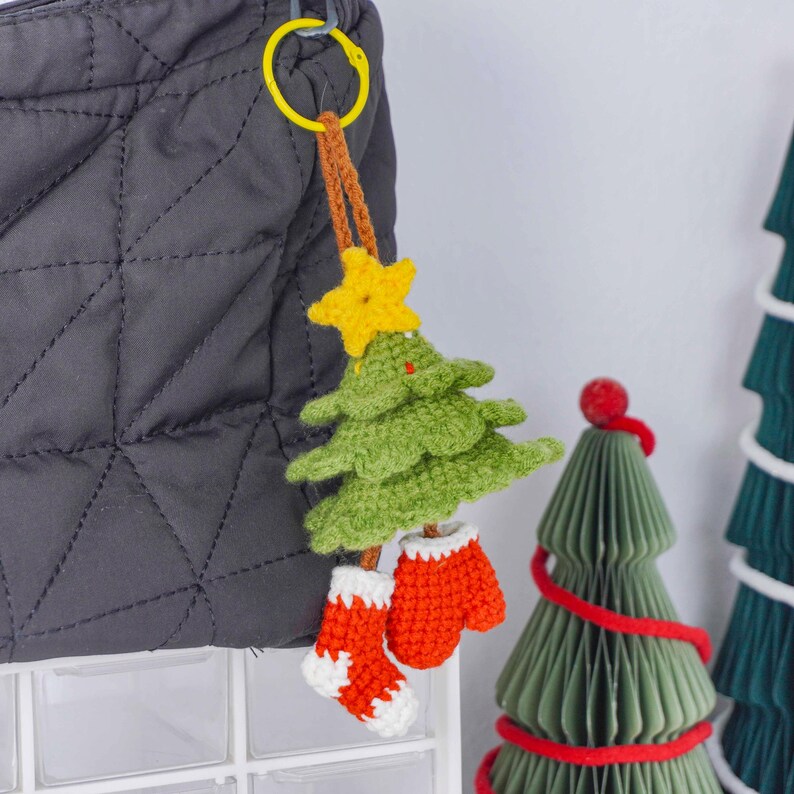 Op de afbeelding: Een handgemaakte kerstboom sleutelhanger met een gele ster, groene boom en rode wanten en sok. De sleutelhanger is bevestigd aan een grijze gewatteerde tas met een gele ring.
