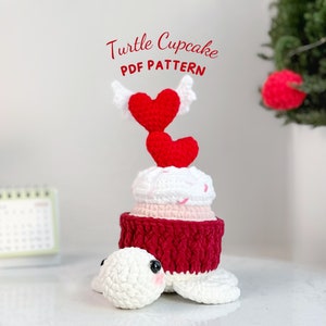 Peut inclure: Jouet amigurumi en crochet en forme de cupcake tortue avec un cœur rouge et blanc sur le dessus. Le cupcake est décoré de glaçage blanc et de pépites roses. La tortue est blanche avec des yeux noirs. Le texte "Turtle Cupcake PDF PATTERN" est écrit au-dessus du jouet.