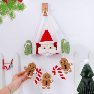 May include: A handmade Christmas decoration featuring a crocheted Santa Claus face with a red hat and white beard. The decoration includes green mittens and a garland of gingerbread men and candy canes. It hangs from a wooden wall mount.