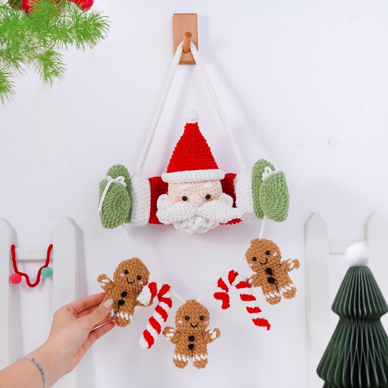 May include: A handmade Christmas decoration featuring a crocheted Santa Claus face with a red hat and white beard. The decoration includes green mittens and a garland of gingerbread men and candy canes. It hangs from a wooden wall mount.