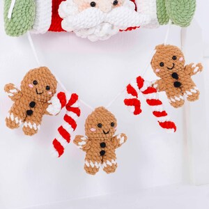 May include: A festive Christmas garland featuring a Santa Claus face and a string of crocheted gingerbread men and candy canes. The gingerbread men are brown with white accents and black button eyes. The candy canes are red and white.