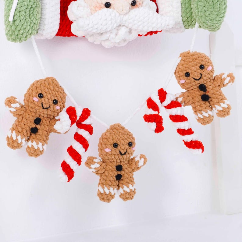 May include: A festive Christmas garland featuring a Santa Claus face and a string of crocheted gingerbread men and candy canes. The gingerbread men are brown with white accents and black button eyes. The candy canes are red and white.