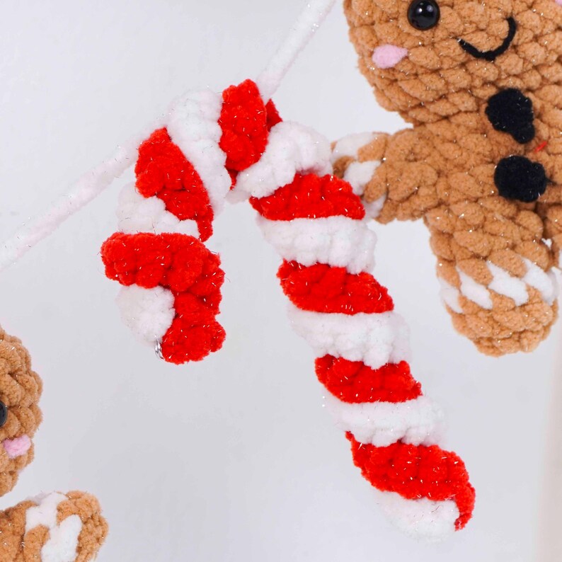 May include: Close-up of a red and white striped candy cane made of soft yarn. The candy cane is part of a Christmas-themed decoration, with a gingerbread man visible in the background. The candy cane is attached to a white, glittery stem.