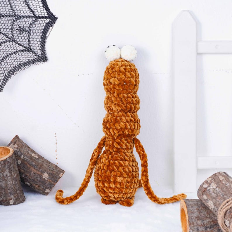 May include: A handmade, plush, brown creature with a segmented body, long arms, and two white pom-pom eyes. The soft, textured toy is likely a Halloween decoration, set against a white backdrop with a spiderweb and wooden accents.