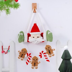 May include: A handmade Christmas wall hanging featuring a crocheted Santa Claus face with a red hat and white beard. The design includes green mittens and a garland of gingerbread men and candy canes, all suspended by a white rope.