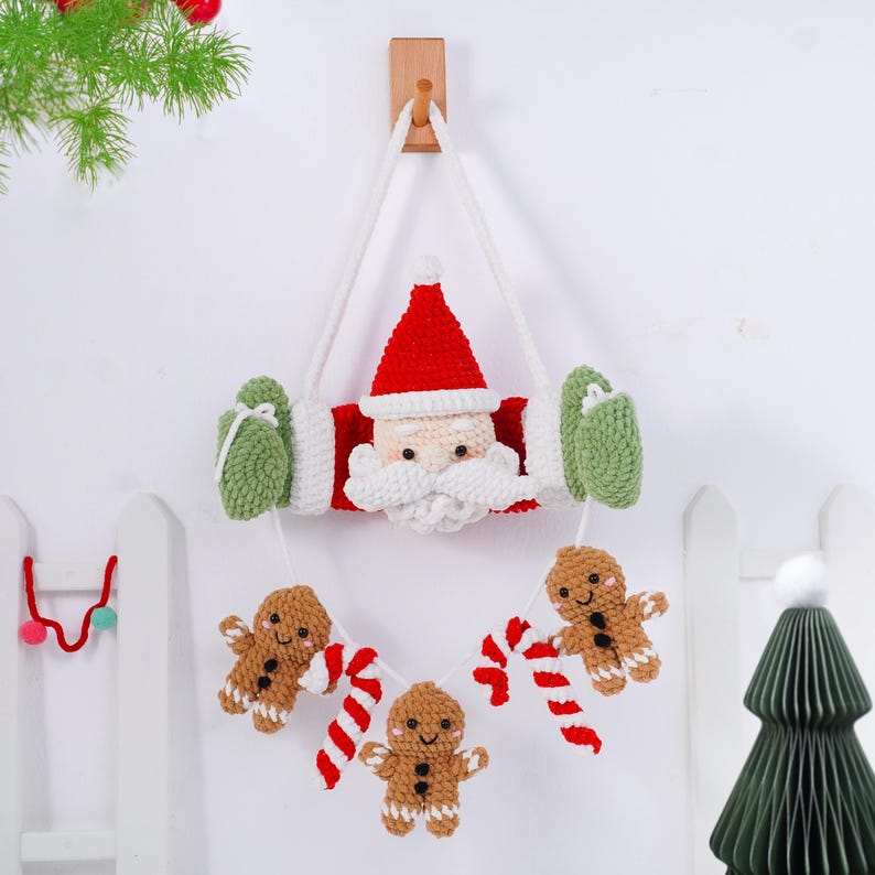 May include: A handmade Christmas wall hanging featuring a crocheted Santa Claus face with a red hat and white beard. The design includes green mittens and a garland of gingerbread men and candy canes, all suspended by a white rope.