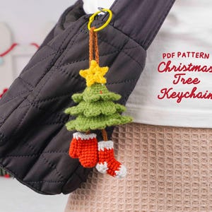 Puede incluir: Un llavero de árbol de Navidad hecho a mano con una estrella amarilla, un árbol verde y mitones y calcetines rojos. El llavero está sujeto a una bolsa acolchada negra. El texto en la camisa dice "PDF PATTERN Christmas Tree Keychain."