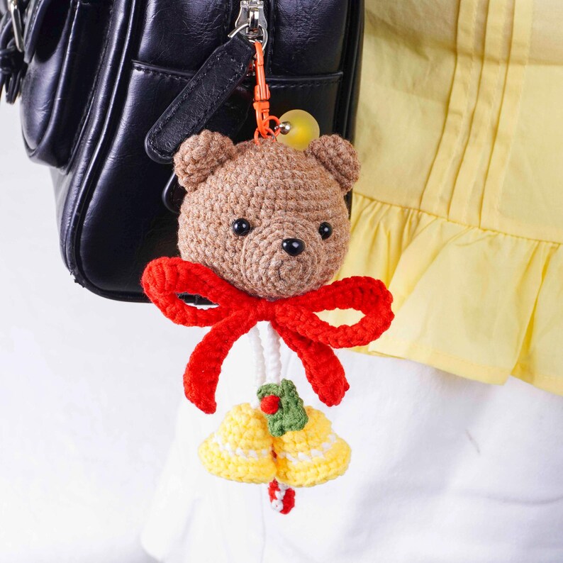 May include: A crocheted teddy bear keychain with a red bow and yellow bells. The bear is brown with black eyes and a stitched nose. The bells are yellow with white accents and a green holly sprig.