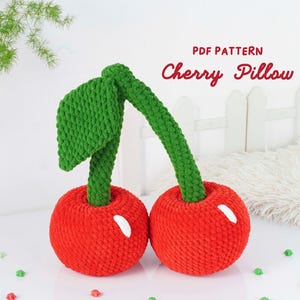 May include: A handmade, plush cherry pillow with a vibrant red body, green stem, and leaf. The pillow has white accents and is labeled "Cherry Pillow" in red script. The pillow is a soft, decorative item.