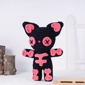May include: A handmade black crochet cat with pink accents. The cat has pink ears, eyes, paws, and a skeleton design on its chest and legs. The eyes are embroidered with an X shape. The cat is standing upright.