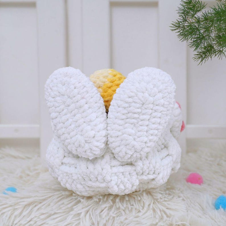 May include: White crocheted bunny basket with yellow and white yarn flower accents. The basket is sitting on a white fluffy surface.