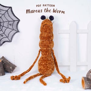 May include: An orange plush worm toy with the text "Marcus the Worm" above it. The worm has a segmented body, long arms and legs, and two dark blue pom-poms on its head. A decorative spiderweb and wooden logs are in the background.