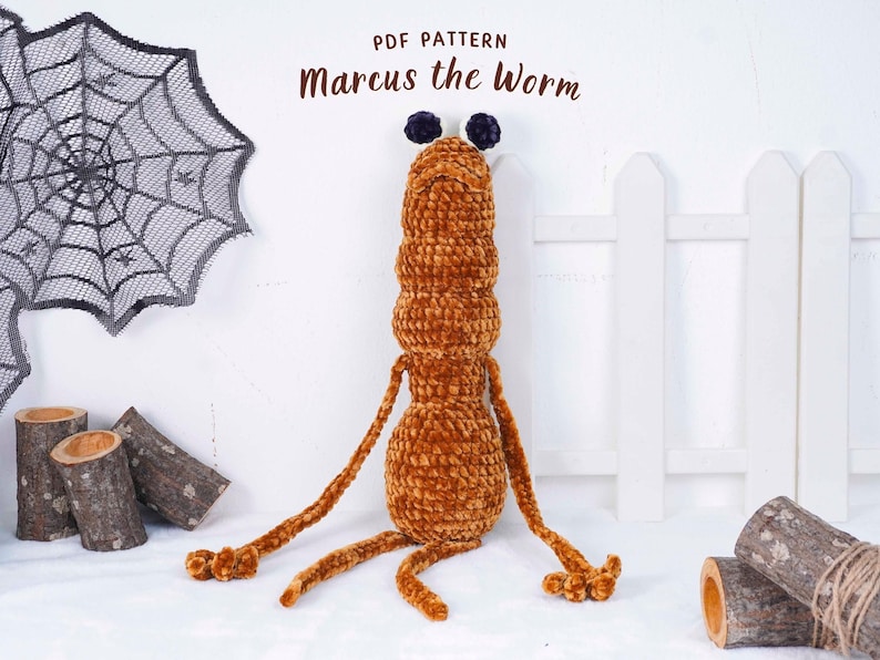May include: A plush, orange worm toy with the text "Marcus the Worm" above it. The worm has a segmented body, long arms and legs, and two dark blue pom-poms on its head. A decorative spiderweb and wooden logs are in the background.