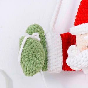 May include: A close-up of a handmade Christmas ornament featuring Santa Claus. The ornament is made of red, white, and green yarn. Santa's hat is red, and his beard and trim are white. He is holding a green gift bag with a white ribbon.