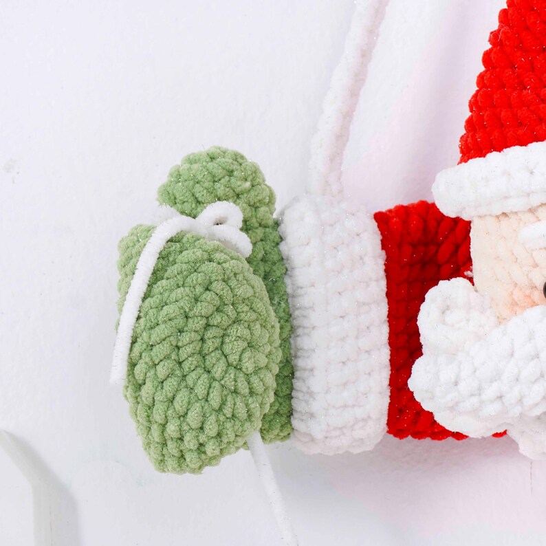 May include: A close-up of a handmade Christmas ornament featuring Santa Claus. The ornament is made of red, white, and green yarn. Santa's hat is red, and his beard and trim are white. He is holding a green gift bag with a white ribbon.