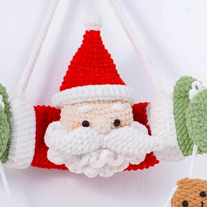 May include: A handmade Christmas ornament featuring a crocheted Santa Claus design. The ornament has a red hat and coat, a white beard and trim, and green mittens. The Santa Claus face is beige with black eyes. White string is attached for hanging.