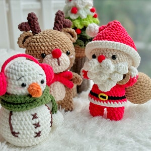 May include: Crocheted Christmas ornaments, including a snowman with a green scarf and red earmuffs, a reindeer with brown fur and red antlers, and a Santa Claus with a white beard and red suit.