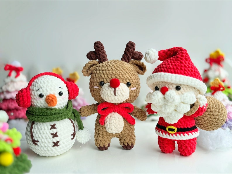 May include: Three crocheted Christmas ornaments: a snowman with a green scarf and red earmuffs, a reindeer with a red bow, and Santa Claus with a white beard and a red bag.
