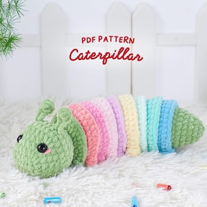 Può includere: Un giocattolo a forma di bruco all'uncinetto fatto a mano nei toni del verde, rosa, giallo e blu. Il bruco ha un aspetto strutturato e morbido con occhi neri e la scritta "PDF PATTERN Caterpillar". Il giocattolo è esposto su una superficie bianca e soffice.
