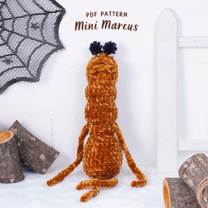 May include: A plush, brown, handmade creature with a segmented body, long arms, and legs. The text "PDF PATTERN Mini Marcus" is at the top. The background includes a white fence, a spider web, and wooden logs.