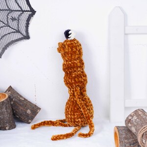 May include: A handmade, plush, brown creature with a small, round head and long, slender legs. The creature has a textured, velvety appearance and a small, white and navy blue ball on top of its head. The creature is sitting on a white surface.
