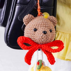 May include: A crocheted teddy bear keychain with a red bow, attached to a black purse. The bear is light brown with black eyes and a black nose. The keychain has a yellow bead and a white beaded strand with a small yellow and green decoration.