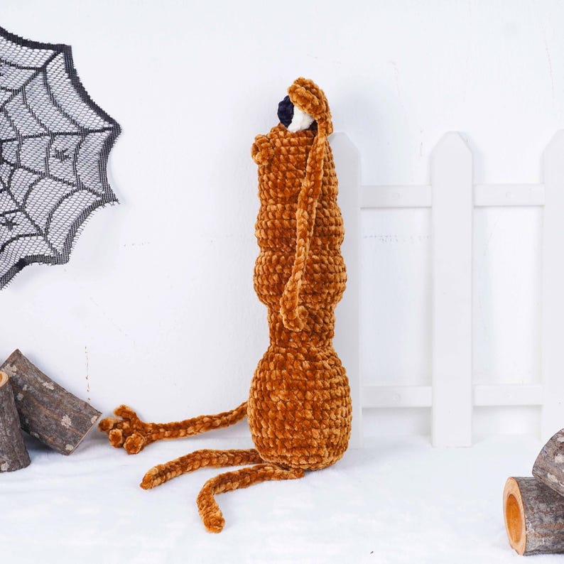 May include: A handmade, brown plush creature with long ears and legs, possibly a dog or rabbit. The plush toy has a textured, velvety appearance and a small, dark eye. The background includes a decorative spiderweb and a white picket fence, suggesting a Halloween theme.
