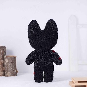May include: A handmade black plush toy with a cat-like head and small arms and legs. The plush has red stitching details on the body. The toy is made of a sparkly, textured yarn.