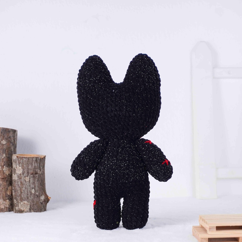 May include: A handmade black plush toy with a cat-like head and small arms and legs. The plush has red stitching details on the body. The toy is made of a sparkly, textured yarn.