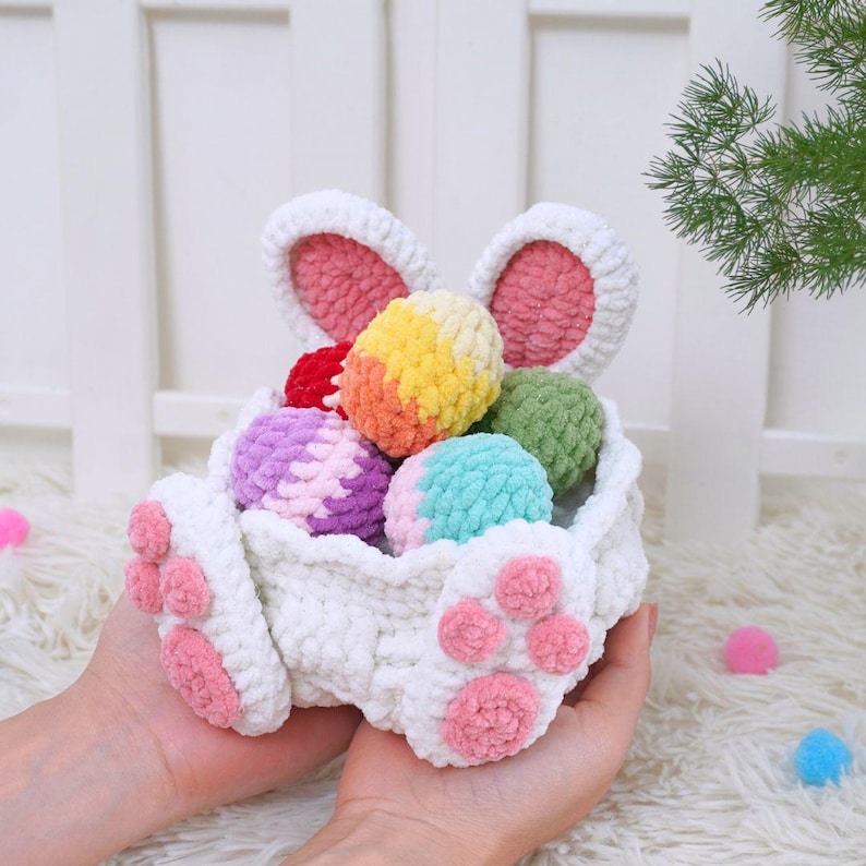 May include: White crocheted bunny basket with pink paw details, filled with colorful yarn Easter eggs.