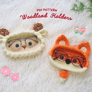 Può includere: Due portaocchiali in crochet a forma di animale. Uno è una pecora bianca con corna marroni e l'altro è una volpe arancione. Entrambi hanno occhiali neri all'interno.