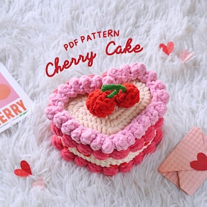 Op de afbeelding: Een gehaakte hartvormige kersentaart tissue box cover. De hoes is roze en wit met een rode kers bovenop. De tekst "PDF PATTERN Cherry Cake" is in zwart op een witte achtergrond geschreven.