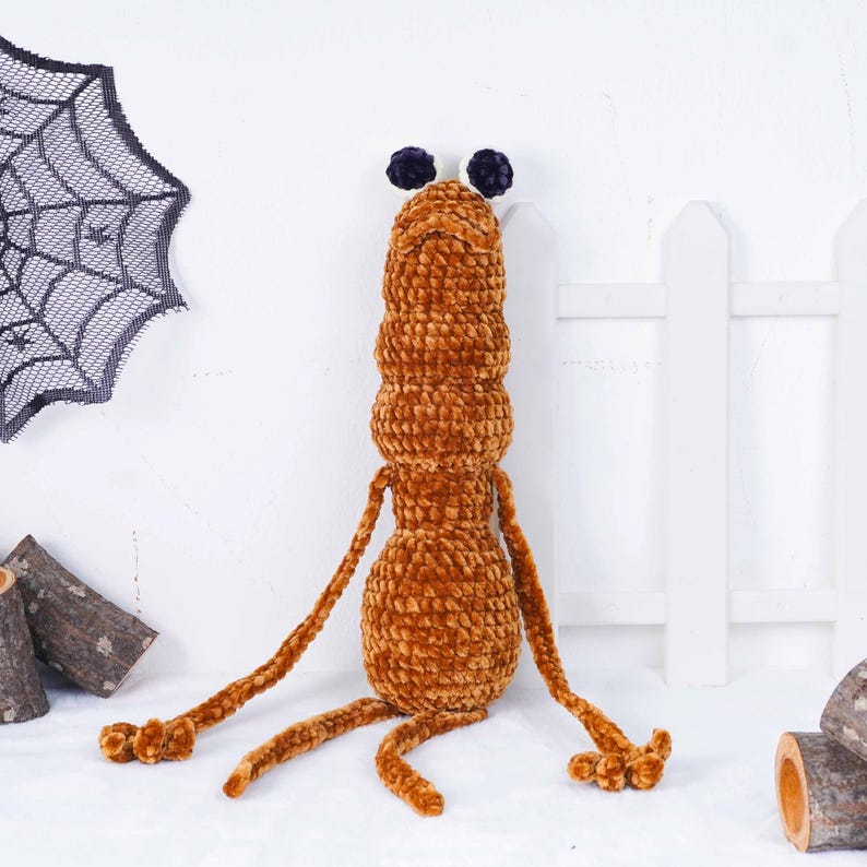 May include: A handmade, plush, brown creature with a segmented body, long arms, and three legs. It has two dark purple, fuzzy eyes. A black spiderweb decoration hangs in the background. This unique plush toy is perfect for Halloween.
