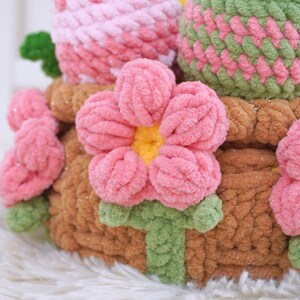 May include: A crocheted flower arrangement in a brown basket. The flowers are pink with yellow centers and green leaves. The basket is decorated with glitter.