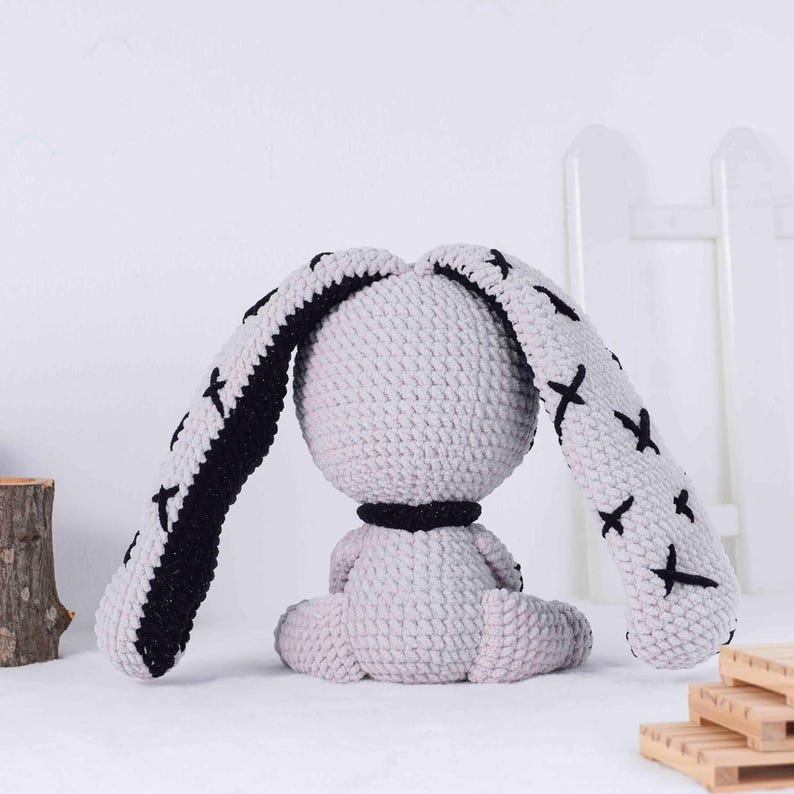 May include: A handmade crochet bunny doll in light gray with black accents. The bunny has long ears with black edges and black cross stitch details. It has a black collar and is sitting down.
