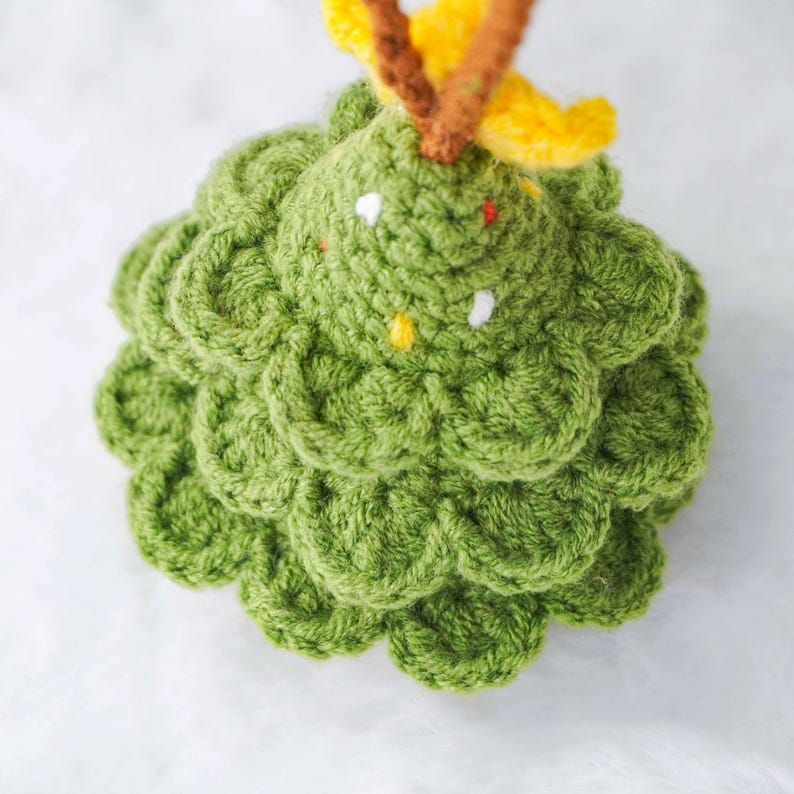 Op de afbeelding: Een handgemaakte, groene gehaakte kerstboomversiering. De boom heeft een getextureerd, gelaagd ontwerp met kleine witte en gele accenten. Het heeft een gele en bruine top en wordt weergegeven tegen een zachte, witte achtergrond.