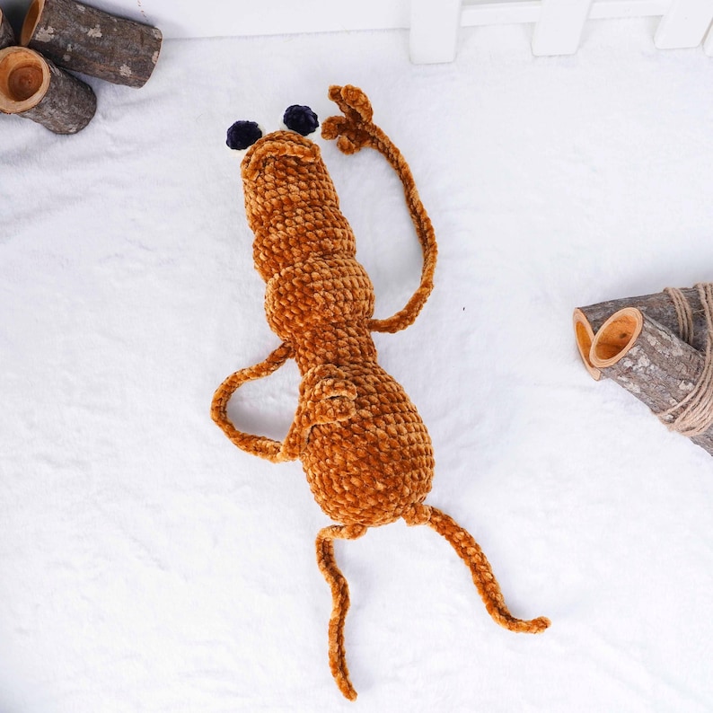 May include: A brown crocheted toy creature with long, thin limbs and two small, dark blue pom-poms on its head. The plush toy is on a white surface, with wooden log accents in the background. The toy has a whimsical, playful design.
