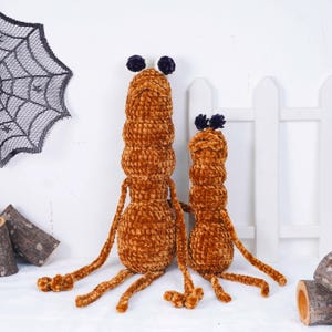 May include: Two brown, plush, worm-like toys with long limbs and small, dark purple antennae. The larger one is taller than the smaller one. A decorative spiderweb and a white picket fence are in the background.