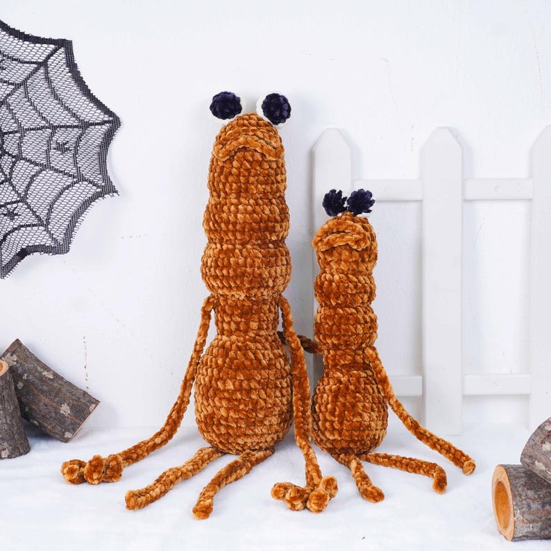 May include: Two brown, plush, worm-like toys with long limbs and small, dark purple antennae. The larger one is taller than the smaller one. A decorative spiderweb and a white picket fence are in the background.