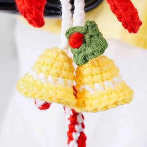 May include: Close-up of a handmade crochet Christmas ornament. The ornament features two yellow bells with white accents, a green leaf with a red berry, and red and white striped accents. The ornament is made of yarn.