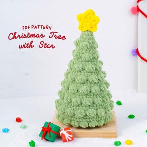 Peut inclure: Un sapin de Noël vert en crochet fait main, surmonté d'une étoile jaune. Le sapin est posé sur une base en bois, avec de petits coffrets cadeaux et des confettis en forme d'étoiles colorées. Le texte sur l'image indique "PDF Pattern Christmas Tree with Star."