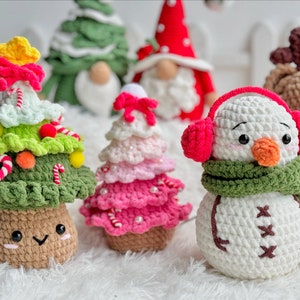 May include: A collection of crocheted Christmas ornaments, including a snowman with a green scarf and red earmuffs, a pink and white Christmas tree, and a green Christmas tree with candy canes and red and yellow ornaments.