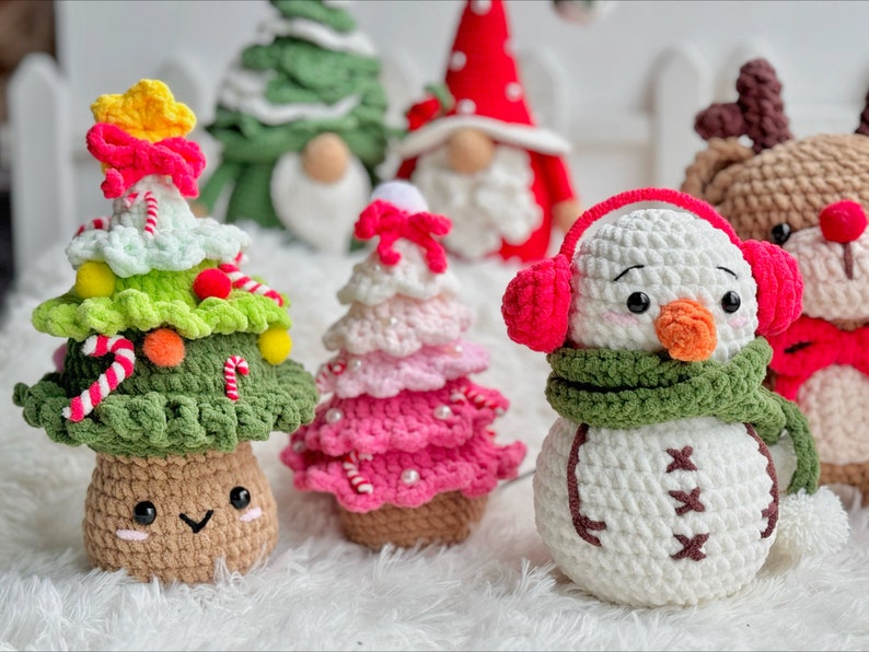 May include: A collection of crocheted Christmas ornaments, including a snowman with a green scarf and red earmuffs, a pink and white Christmas tree, and a green Christmas tree with candy canes and red and yellow ornaments.